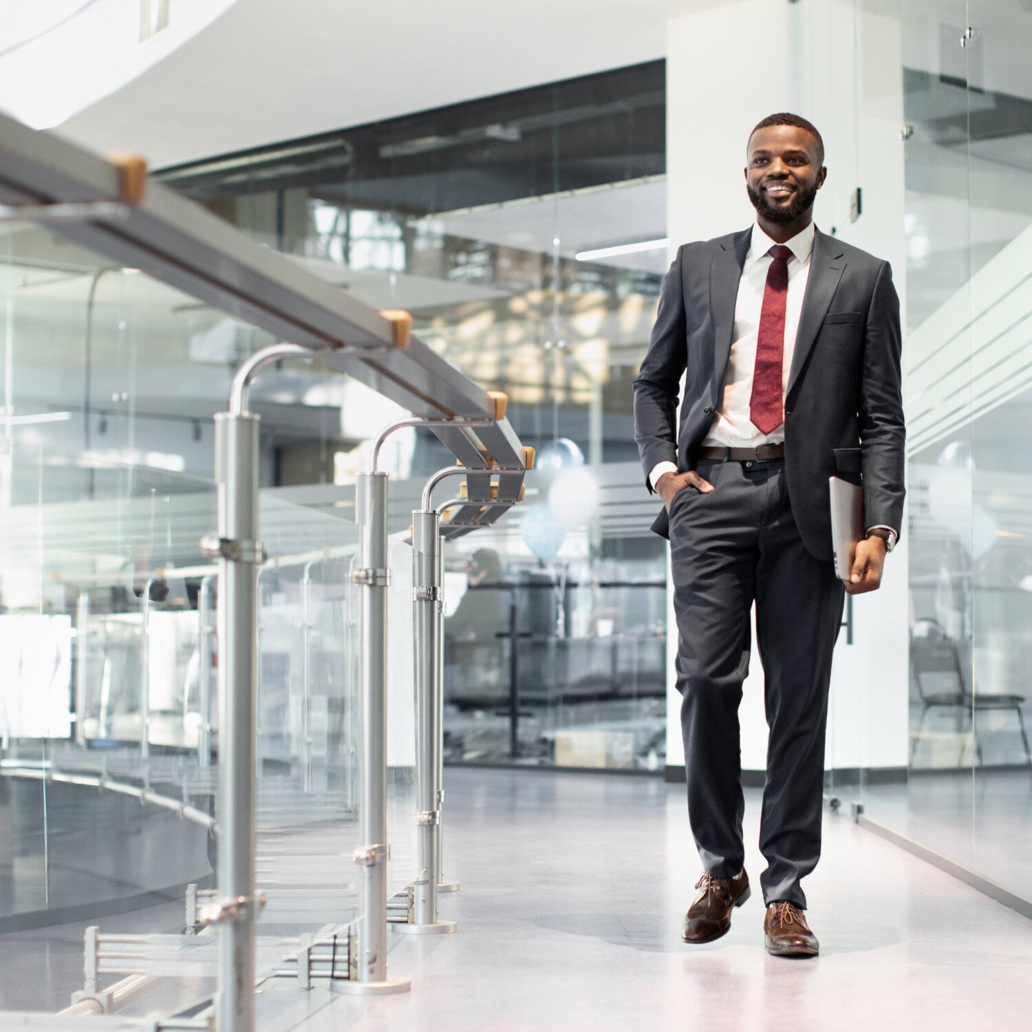 Positive young black manager in suit walking by office building, carrying laptop, looking at copy space and smiling, going to conference with colleagues, panorama, full length shot